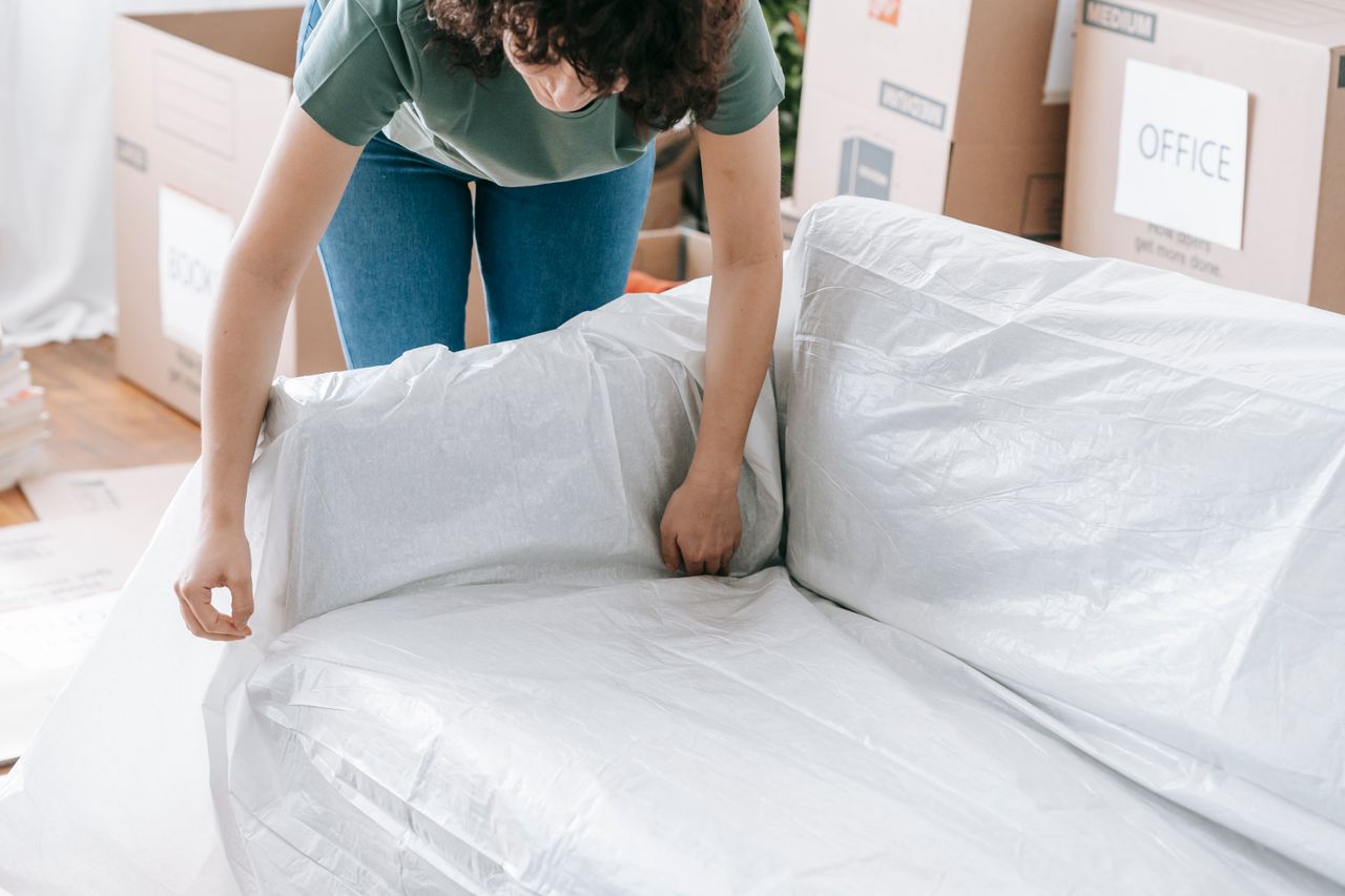 hero-img-02 A woman carefully wraps a sofa in protective material while packing boxes in a bright room.